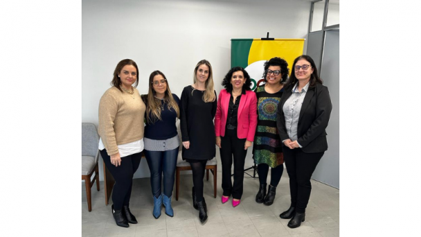 Pessoas em pé, na entrada da Ouvidoria-Geral do Estado. 
Dá esquerda para a direita, Analista Jurídica Bárbara Zucchetti está utilizando blusa cor caramelo de manga longa e calça preta e uma bota preta com pouco salto.

Ao seu lado, Carolina Vendruscolo Chefe da Divisão de Manifestações está utilizando uma blusa preta de manga longa e calça azul com uma bota azul de salto médio.

A Ouvidora-Geral do Estado Viviane Migliavacca está utilizando vestido até o joelho, na cor preta, e uma bota preta com pouco salto.

Fátima Anise Rodrigues Ehlert Presidente do Conselho Estadual de Educação está utilizando blusa na cor preta e um blazer rosa pink, uma calça preta e um sapato de bico fino, na cor rosa, com abertura em cima. 

Márcia Chaves Chefe da Divisão de Transparência está utilizando um vestido até o joelho, na cor preta  com mandalas coloridas e de manga longa, e uma meia-calça preta e uma bota preta com pouco salto.

Ouvidora da temática da Educação Jucele Comis está utilizando blusa na cor cinza e um blazer preta, e uma calça preta e uma bota preta com pouco salto.
